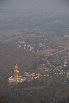 High angle daytime view from the mountain peak at Khao Wong Phrachan in Lopburi Thailand looking down at the giant golden seated Buddha statue and temple complex surrounded by dry forest hills in haze
