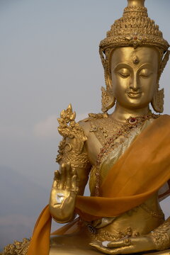 Detailed close vertical view of an ornate golden crowned Buddha statue draped in a saffron robe with raised hand gesture against a soft hazy sky at the summit of Khao Wong Phrachan in Lopburi Thailand