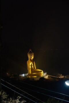 Vertical night view from the mountain trail at Khao Wong Phrachan in Lopburi Thailand showing the giant golden seated Buddha statue glowing against the dark sky with lit temple shrine and stairs below