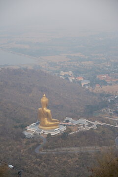High angle daytime view from the mountain peak at Khao Wong Phrachan in Lopburi Thailand looking down at the giant golden seated Buddha statue and temple complex surrounded by dry forest hills in haze