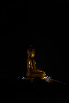 Side profile view of a giant golden seated Buddha statue illuminated at night against the pitch black sky captured from the way up the mountain trail at Khao Wong Phrachan in Lopburi province Thailand