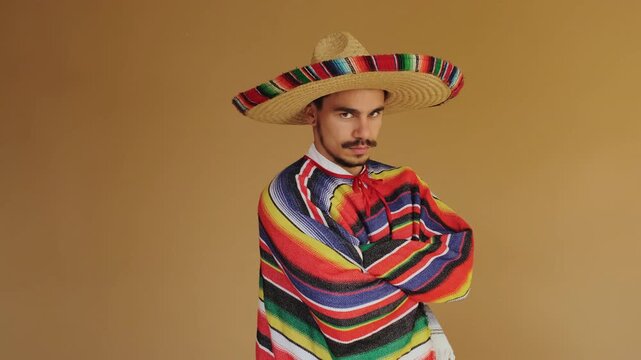 Adult male wearing large woven straw sombrero with multicolor trim and bright striped serape, positioned against warm tan studio backdrop while gaze rises from downcast to confident portrait
