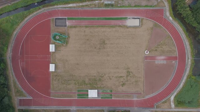 Top down aerial drone footage slowly zooming in toward an outdoor athletics running track. Sports facility with red oval track, field areas, lanes, greenery and surrounding water viewed from directly 