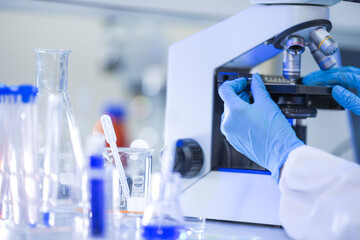 A female laboratory technician in a white coat, protective eyewear and a sterile hairnet twisting objective turret toward higher magnification inside a busy medical diagnostics laboratory. © ultramansk