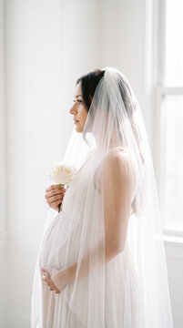 Pregnant woman in white dress with bridal veil holding rose in profile portrait