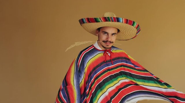 Young man in woven straw sombrero with bright woven band and striped serape poses against warm tan background, holding reed near mouth while making playful expressive hand gestures