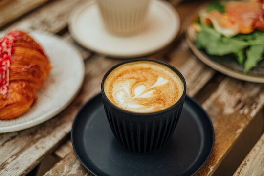 Close-up of a creamy latte in a black cup, surrounded by a croissant and savory brunch dishes on a weathered wooden caf&eacute; table