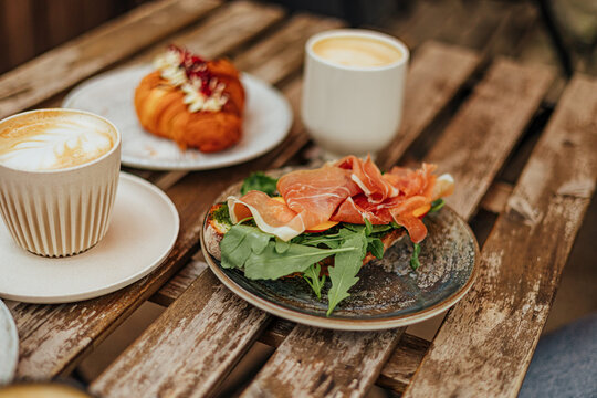 Open toast topped with arugula and thin slices of cured ham, served with two creamy coffees and a pastry on a weathered wooden caf&eacute; table