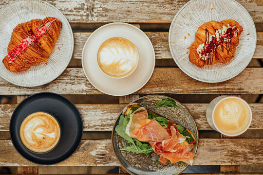Overhead caf&eacute; table with three coffees, decorated croissants, and an open sandwich topped with arugula and cured ham on rustic wooden slats