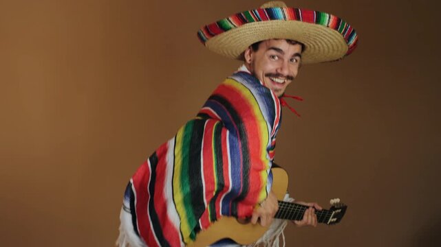 Young man wearing woven straw sombrero and brightly striped poncho turns torso and head toward camera while holding acoustic guitar, smiling broadly with visible teeth and raised eyebrows to convey