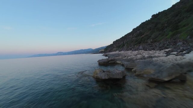 Proximity flying FPV drone footage skimming the calm sea along a rocky shoreline at dusk, pink evening sky over Cala Gonone, Sardinia, Italy.