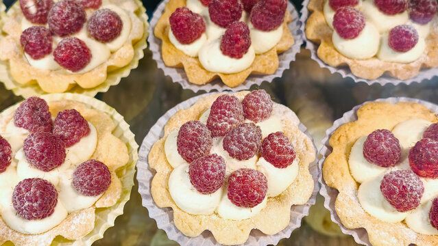 Raspberry tarts with creamy white filling and fresh red berries, displayed in a bakery window, showcasing sweet dessert pastries for sale