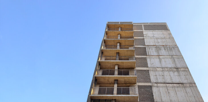 Unfinished residential tower with multiple balconies showing raw concrete and exposed brickwork, symbolizing urban development, progress, and future housing projects against a clear blue sky