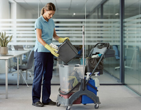 A female cleaner in yellow gloves emptying a trash bin into a plastic bucket on a janitor trolley in a modern office.