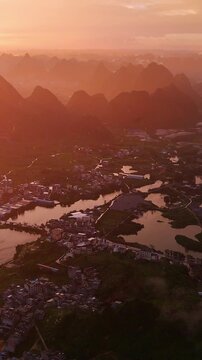 Vertical Video: Golden sunlight breaking through rain over a riverside village with karst mountains in the distance, creating a misty glow