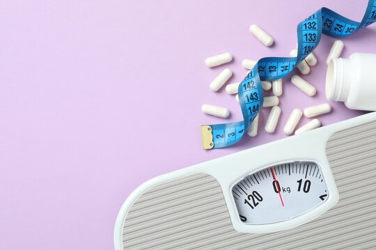 Scales with weight loss pills and measuring tape on violet background, top view