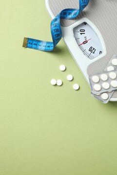 Scales with weight loss pills and measuring tape on green background, top vertical view