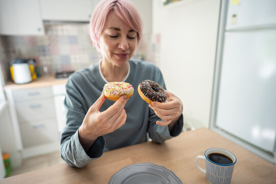 Smiling mature woman holds two colorful donuts after giving in to intense PMS cravings, showing dietary relapse, sugar addiction and return to sweet pastries despite previous effort to quit sweets.