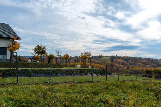 Modern private garden with trimmed lawn, ornamental trees and metal fencing overlooks rolling countryside covered in golden autumn foliage. Wide rural landscape under dramatic clouds suits landscaping