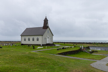Fototapeta premium Strandarkirkja stands alone in low coastal grass at Selvogur, Iceland, beneath an expansive stormy sky. The white wooden church occupies the low horizon, emphasizing the scale of the open plain.