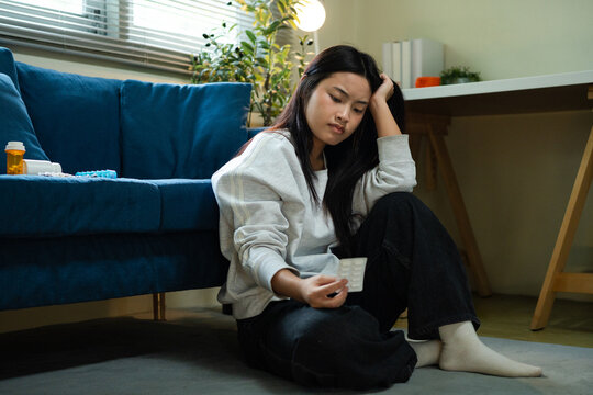 Depressed Young Asian Woman Sitting on Floor Holding Cup with Medication Nearby in Living Room