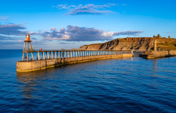 Historic pier and navigation beacon at Whitby Harbour with the dramatic East Cliff and ruins of Whitby Abbey in warm evening sunlight, North Yorkshire, England.