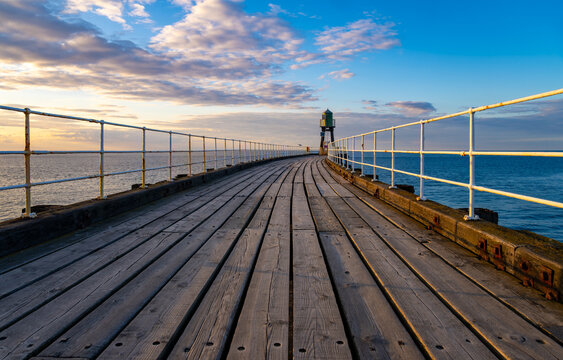 Wooden pier at Whitby Harbour leading towards a navigation beacon in warm evening light, with dramatic clouds over the North Sea in North Yorkshire, England.
