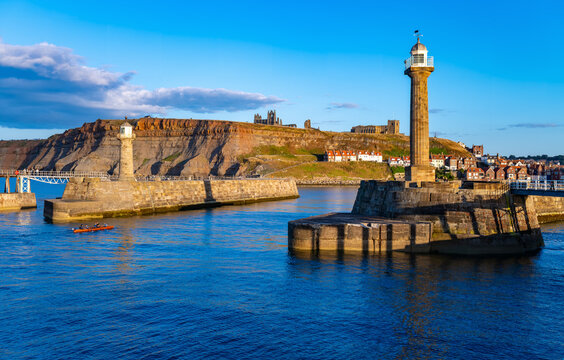 Historic lighthouses at the entrance to Whitby Harbour with the ruins of Whitby Abbey on the East Cliff, illuminated by warm evening light in North Yorkshire, England.