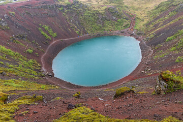 A broad crater basin under low rain cloud appears at Kerid Crater, Iceland. Diffuse afternoon light reveals turquoise water ringed by red and green volcanic terrain around the volcanic basin. © Artem