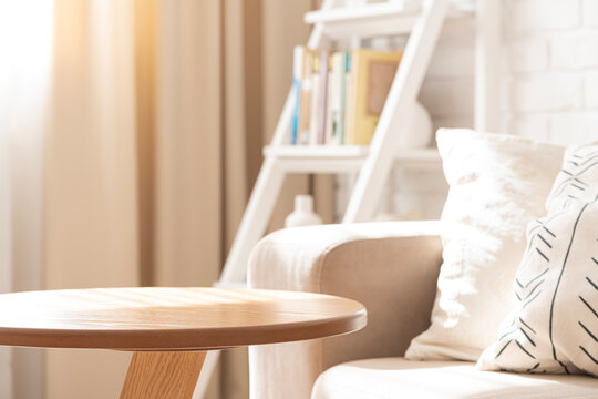 Empty round wooden table in dappled sunlight near sofa in blurred room interior background