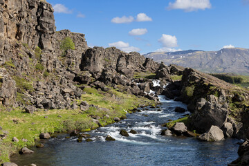 Fototapeta premium Oxara River flows past angular rocks beneath open blue sky in Thingvellir National Park, Iceland. A clear channel leaves the narrow gorge. Water guides the eye toward sunlit terrain.