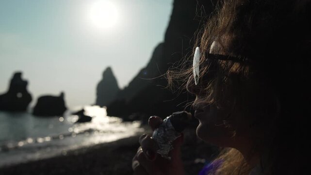 Sunset beach eating, woman enjoys snack against ocean cliffs and bright sun during summer vacation travel