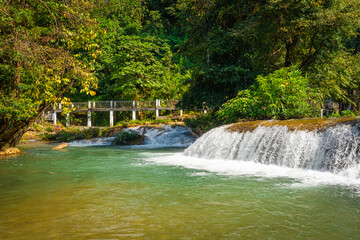 Scenic view of clear stream flowing over small rock cascades with a white bridge amidst dense green jungle foliage at Glue-Thor Waterfall (Kyone Htaw Waterfall), Kayin State, Myanmar. © Around Ball
