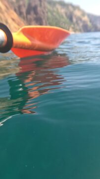 Kayaking paddle sea shot of an orange blade hovering over clear turquoise water near coastal cliffs during a peaceful summer adventure trip
