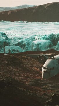A plane wreck sits on rocky ground beside a large glacier. Ice towers rise in the background, showing a cold and isolated environment. The landscape appears harsh and uninviting.