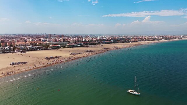 High aerial view of La Malvarrosa beach in Valencia, Spain. Panoramic shot showing the crowded sandy coastline, city skyline, Mediterranean sea with a sailboat, and the port in the distance under a su