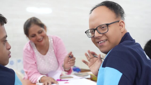 Indian disabled student with down syndrome smiling and waving at the camera while sitting with a teacher in a special needs classroom, representing inclusive education and learning