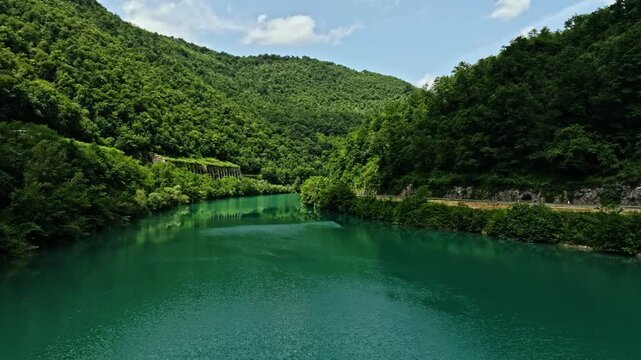 Mavic 3 Pro drone aerial forward flight along the turquoise Soca river in Slovenia. Scenic landscape view with a mountain road visible on the right side and green hills on the distance horizon.