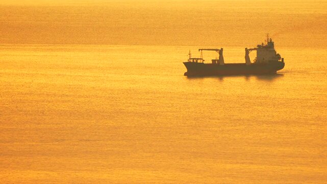 Cargo ship, golden ocean, dramatic sunset silhouette of vessel cruising across the glowing sea water during beautiful sunrise or dusk.