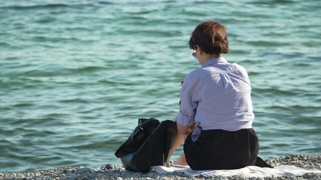 Woman Beach Ocean. Woman sitting on pebble beach looking at the ocean, using phone, enjoying leisure.
