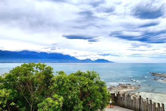 Kaikoura, New Zealand - Coastal Lookout Over Blue Sea And Mountain Range, Framed By A Green Tree And Wooden Railing Under A Cloudy Sky, Capturing The Region&rsquo;s Signature Blend Of Rugged Shoreline.