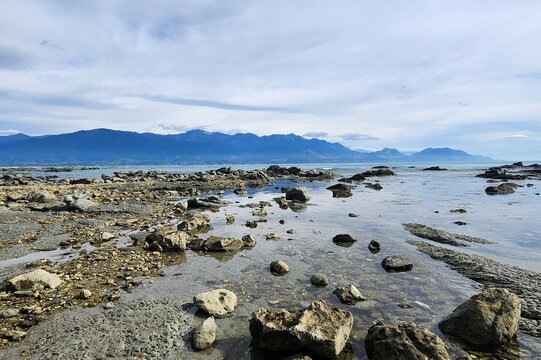 Kaikoura, New Zealand -  Rocky Shoreline With Shallow Tide Pools Reflecting The Sky, Overlooking Blue Coastal Water And A Distant Mountain Range Under A Partly Cloudy Atmosphere.