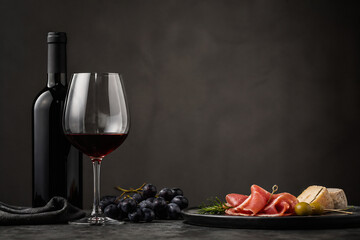 Naklejka premium A bottle of red wine and a glass of red wine are displayed next to a platter of sliced cured meats, grapes, and cheese on a dark surface against a muted background.