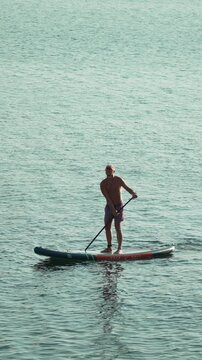 Paddleboard man water, a fit man paddleboarding alone on calm clear sea water enjoying summer healthy outdoor activity. Vertical video