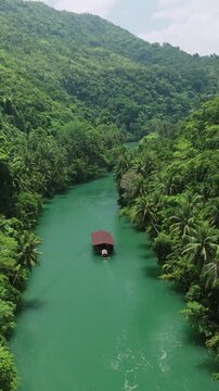 Vertical drone tracks and follows a Loboc River cruise boat downstream through green water between forested hills in Bohol