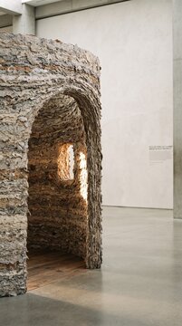Vertical Sustainable art installation with a rough recycled paper texture showing a sculptural hut with warm interior light inside a minimalist concrete museum gallery, representing eco-friendly mater