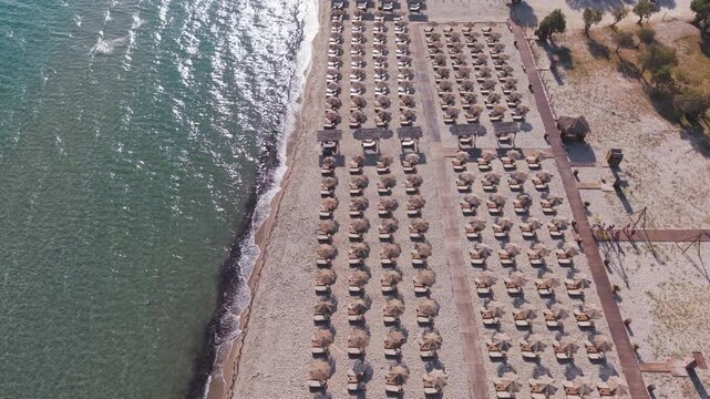 Wide aerial view of a developed sandy beach with numerous umbrellas and sun loungers arranged in rows beside clear coastal water and pedestrian walkways.