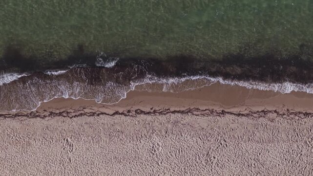 Overhead aerial view of foamy sea waves washing onto an empty sandy shoreline, creating natural textures and parallel coastal patterns.
