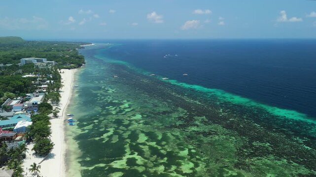Drone pulls back and ascends over Alona Beach, revealing white sand, shallow reef waters, coastline, and deep blue sea in Panglao, Bohol