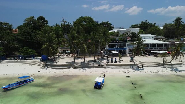 Drone slides right over Alona Beach, revealing white sand, shallow turquoise water, boats, palms, and beachfront buildings in Panglao, Bohol